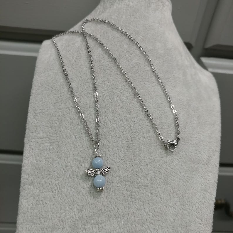 May include: A silver-toned necklace featuring a pendant shaped like an angel. The angel pendant has a light blue bead body and decorative wings. The necklace is displayed on a gray surface.