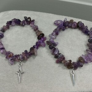 May include: Two amethyst chip bead bracelets. Each bracelet features a mix of purple and brown gemstone chips. One bracelet has a silver star charm, and the other has a silver spike charm. The bracelets are displayed on a light grey surface.