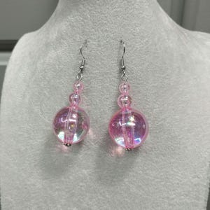 May include: A pair of pink iridescent dangle earrings. Each earring features a large, round, transparent bead at the bottom, with three smaller beads above it. The earrings have silver-colored hooks.