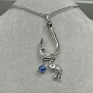 May include: Silver-toned necklace featuring a large fish hook pendant. A small fish charm and a blue bead dangle from the hook. The chain is a curb style. The necklace is displayed against a neutral background.