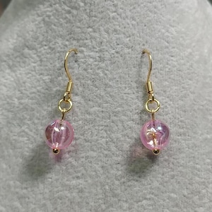 Peut inclure: Une paire de boucles d'oreilles pendantes en or avec des perles de verre roses. Les perles ont une finition chatoyante.