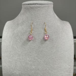 May include: A pair of gold dangle earrings with pink glass beads. The beads are round and have a slightly textured surface.