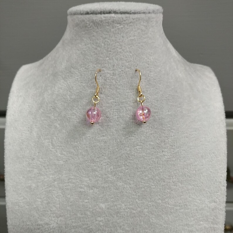 May include: A pair of gold dangle earrings with pink glass beads. The beads are round and have a slightly textured surface.