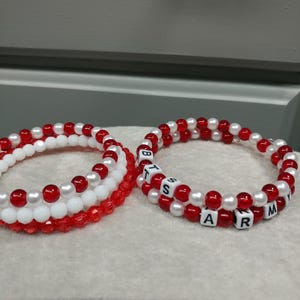 Könnte beinhalten: Zwei Armbänder mit Perlen. Ein Armband hat rote und weiße Perlen. Das andere hat rote, weiße und beschriftete Perlen mit der Aufschrift "BTS ARMY". Die Armbänder liegen auf einer weißen Oberfläche.