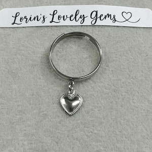 May include: A silver ring with a dangling heart charm. The ring is simple and elegant, and the heart charm adds a touch of whimsy.