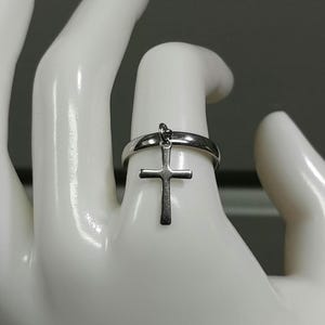 May include: A silver ring featuring a dangling cross pendant. The ring is displayed on a white hand, with the cross suspended from the ring's band. The cross has a simple, classic design.