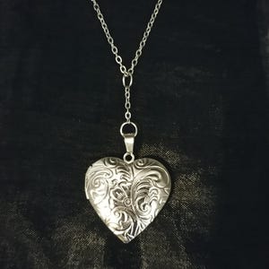 Może przedstawiać: Srebrzysty wisiorek w kształcie serca zawieszony na delikatnym, pasującym metalowym łańcuszku. Powierzchnia serca posiada misterny, tłoczony wzór kwiatowy, który pokrywa cały przód metalowego elementu w kształcie medalionu.