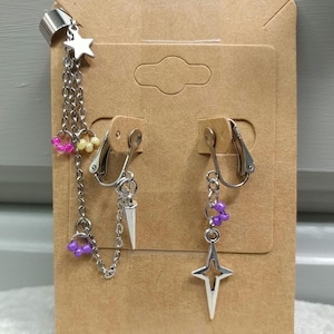 May include: Silver-toned ear cuff and clip-on earrings set. The ear cuff features a star charm and chain with pink, yellow, and purple beads. One clip-on earring has a chain with a silver spike charm, and the other has a star charm with purple beads.