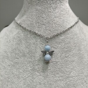 May include: A silver-toned necklace featuring a pendant shaped like an angel. The pendant has two light blue spherical beads and decorative wings. The chain is made of small, interconnected links, and the necklace is displayed against a gray background.