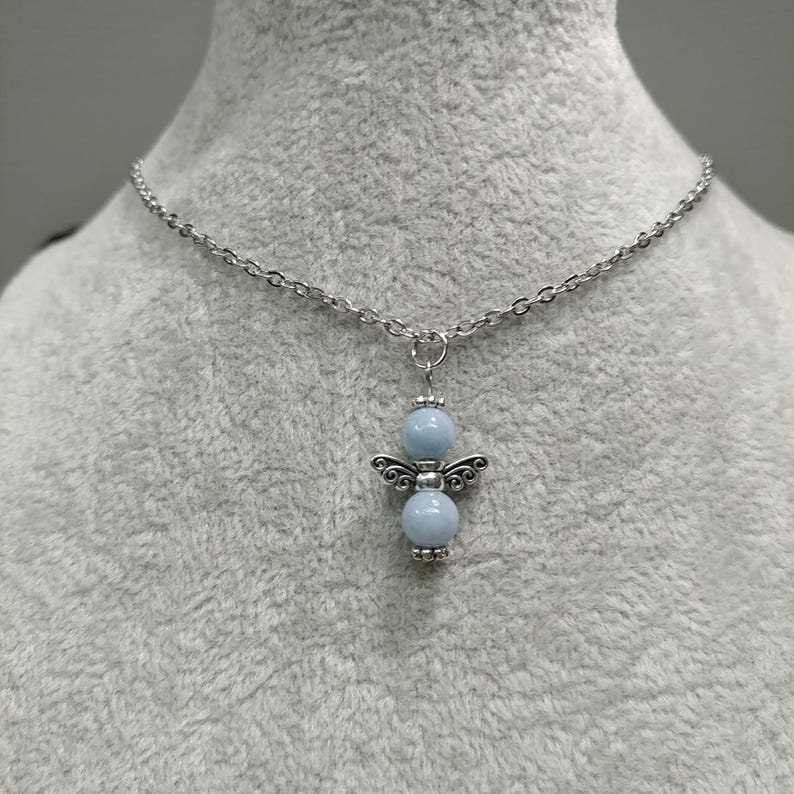May include: A silver-toned necklace featuring a pendant shaped like an angel. The pendant has two light blue spherical beads and decorative wings. The chain is made of small, interconnected links, and the necklace is displayed against a gray background.