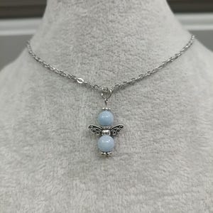 May include: A silver-toned necklace featuring a pendant shaped like an angel. The pendant has two light blue beads and silver wings. The chain is a delicate link style.