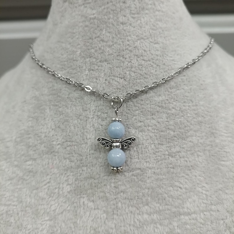 May include: A silver-toned necklace featuring a pendant shaped like an angel. The pendant has two light blue beads and silver wings. The chain is a delicate link style.