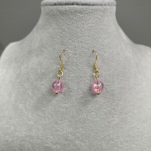 May include: A pair of gold dangle earrings with pink glass beads. The beads have a textured surface and are translucent.