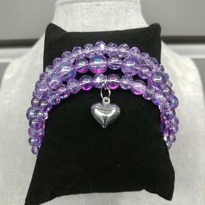 May include: A beaded bracelet with three strands of iridescent purple beads. A silver heart charm dangles from the center. The bracelet is displayed on a black velvet surface.