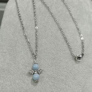 May include: A silver-colored necklace featuring a pendant in the shape of an angel. The angel pendant has a light blue bead for the head and body, with silver-colored wings and decorative details. The necklace chain is also silver-colored.