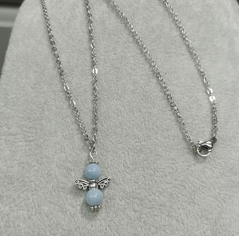 May include: A silver-colored necklace featuring a pendant in the shape of an angel. The angel pendant has a light blue bead for the head and body, with silver-colored wings and decorative details. The necklace chain is also silver-colored.
