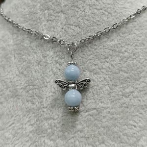 May include: A silver-toned necklace featuring a pendant in the shape of an angel. The angel has light blue bead body and wings. The necklace chain is also silver-toned.