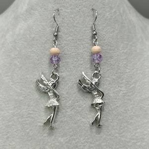 Puede incluir: Un par de pendientes colgantes con dijes de hadas plateadas. Cada pendiente tiene un gancho plateado, una cuenta de madera de color claro, una cuenta púrpura y un dije de hada con alas extendidas. Los pendientes están diseñados para orejas perforadas.