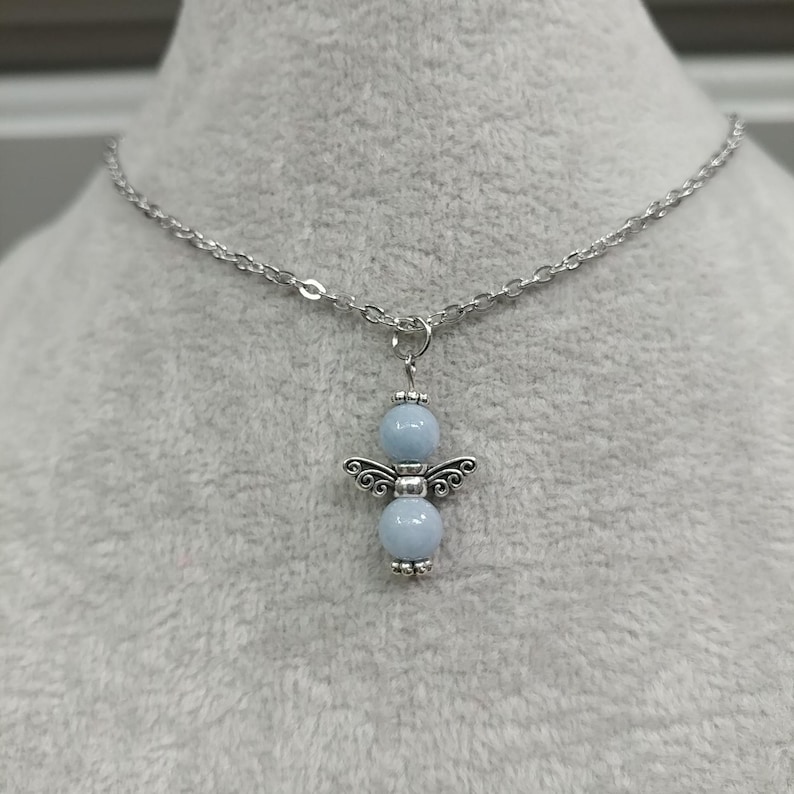 May include: A silver-toned necklace featuring a pendant in the shape of an angel. The angel pendant has a light blue bead for the head and body, with silver-toned wings and feet. The necklace chain is also silver-toned.