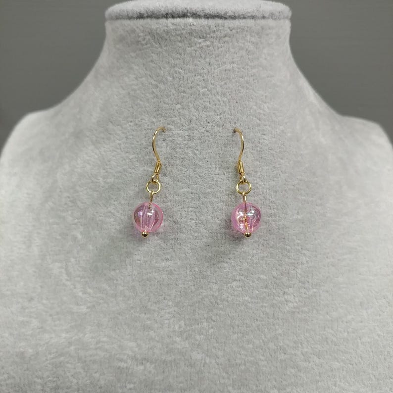 May include: A pair of gold dangle earrings with pink glass beads. The beads are round and have a slight shimmer.