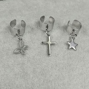 May include: Three silver-toned ear cuffs with dangling charms. One cuff has a butterfly charm, another a cross, and the third a star. The cuffs are open-ended and designed to wrap around the ear.