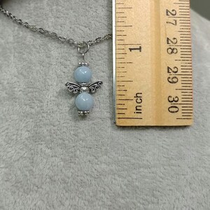 May include: A silver-toned necklace with a pendant shaped like an angel. The angel pendant features light blue beads for the body and decorative silver wings. A wooden ruler is next to the necklace, showing measurements in inches.