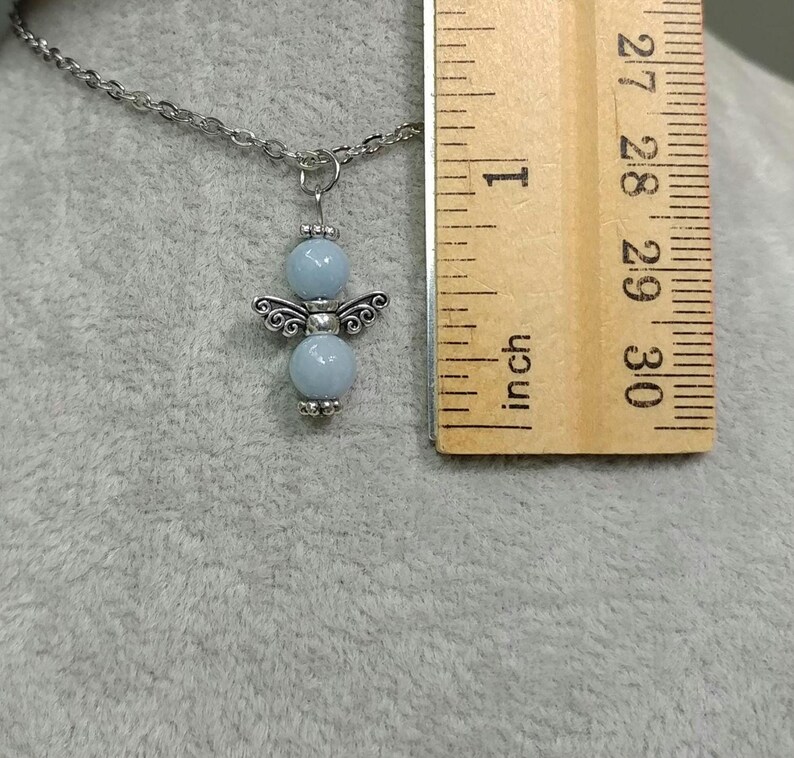 May include: A silver-toned necklace with a pendant shaped like an angel. The angel pendant features light blue beads for the body and decorative silver wings. A wooden ruler is next to the necklace, showing measurements in inches.
