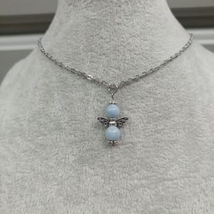 May include: A silver-toned necklace featuring a pendant in the shape of an angel. The angel pendant has two light blue beads for the body and head, with decorative silver wings. The necklace chain is also silver.