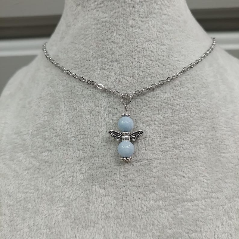 May include: A silver-toned necklace featuring a pendant in the shape of an angel. The angel pendant has two light blue beads for the body and head, with decorative silver wings. The necklace chain is also silver.