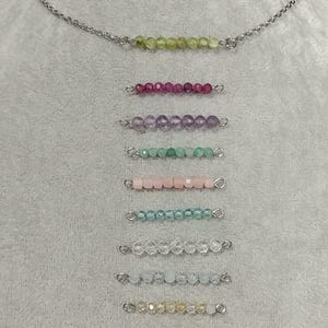 Puede incluir: Un collar de cadena de plata con 11 hebras de cuentas de piedras preciosas de diferentes colores. Cada hebra tiene un cierre de plata en cada extremo. Las hebras están dispuestas verticalmente, con la hebra verde en la parte superior y la hebra morada en la parte inferior.