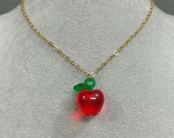 Puede incluir: Un collar de cadena dorada con un colgante en forma de manzana roja. La manzana tiene una hoja y un tallo verdes. El collar se muestra sobre un fondo gris claro.
