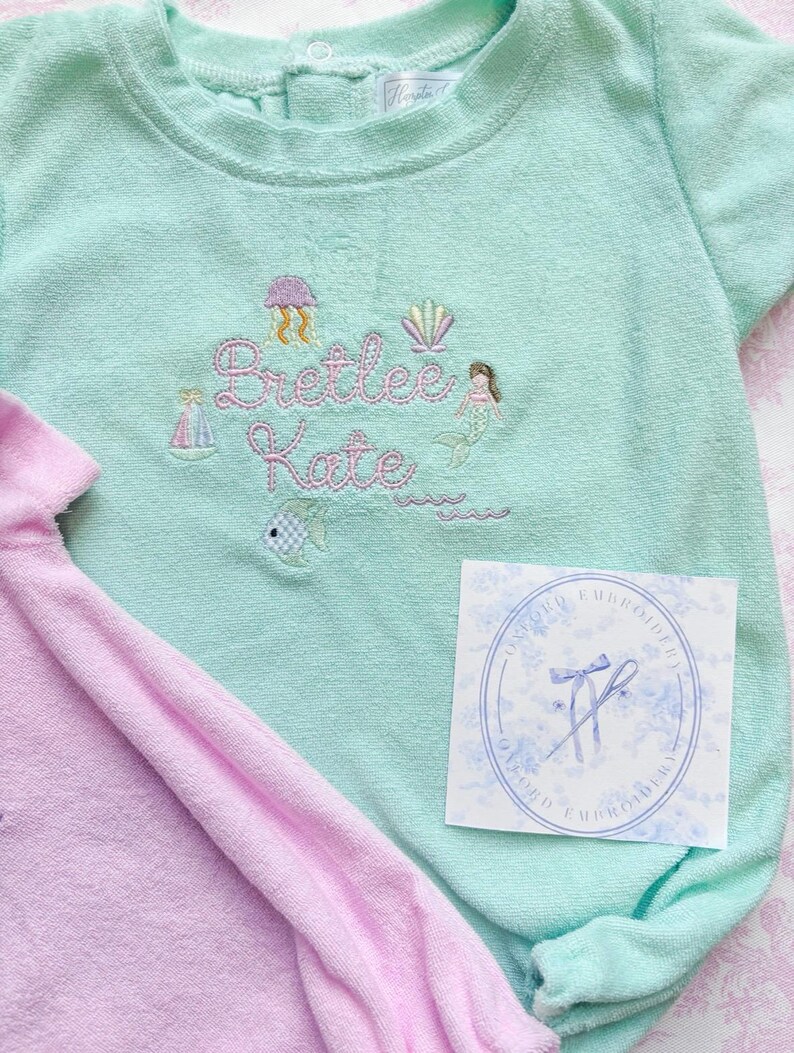 May include: A light teal baby romper with embroidered details, including a jellyfish, seashell, mermaid, sailboat, and the name "Bretlee Kate." A pink romper is partially visible. A white tag with a blue floral design is also present.