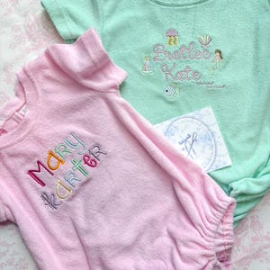 May include: Two baby outfits, one pink and one mint green, with embroidered names and nautical-themed designs. The pink outfit has the name "Mary Karter" embroidered in colorful thread. The green outfit features the name "Bretlee Kate" with a mermaid and other sea creatures.