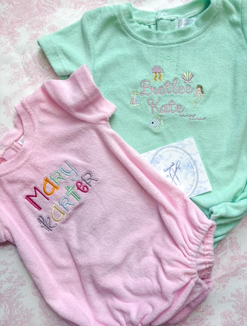 May include: Two baby outfits, one pink and one mint green, with embroidered names and nautical-themed designs. The pink outfit has the name "Mary Karter" embroidered in colorful thread. The green outfit features the name "Bretlee Kate" with a mermaid and other sea creatures.