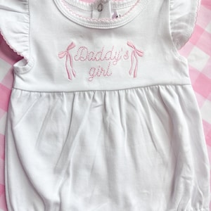 May include: White baby romper with pink trim and flutter sleeves. The romper features the embroidered text "Daddy's girl" in pink script, with pink bow accents on either side. The garment is made of soft fabric and has a snap closure.