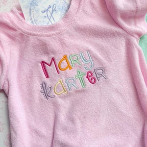 May include: A light pink terry cloth romper with short sleeves. The name "MARY KARTER" is embroidered in colorful thread across the chest. A tag with a blue floral design is visible near the neckline. The romper is designed for babies.