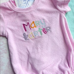 May include: A light pink terry cloth baby romper with the name "Mary Karter" embroidered in colorful thread. The romper has short sleeves and a round neckline. A small card with a logo is tucked into the neckline.