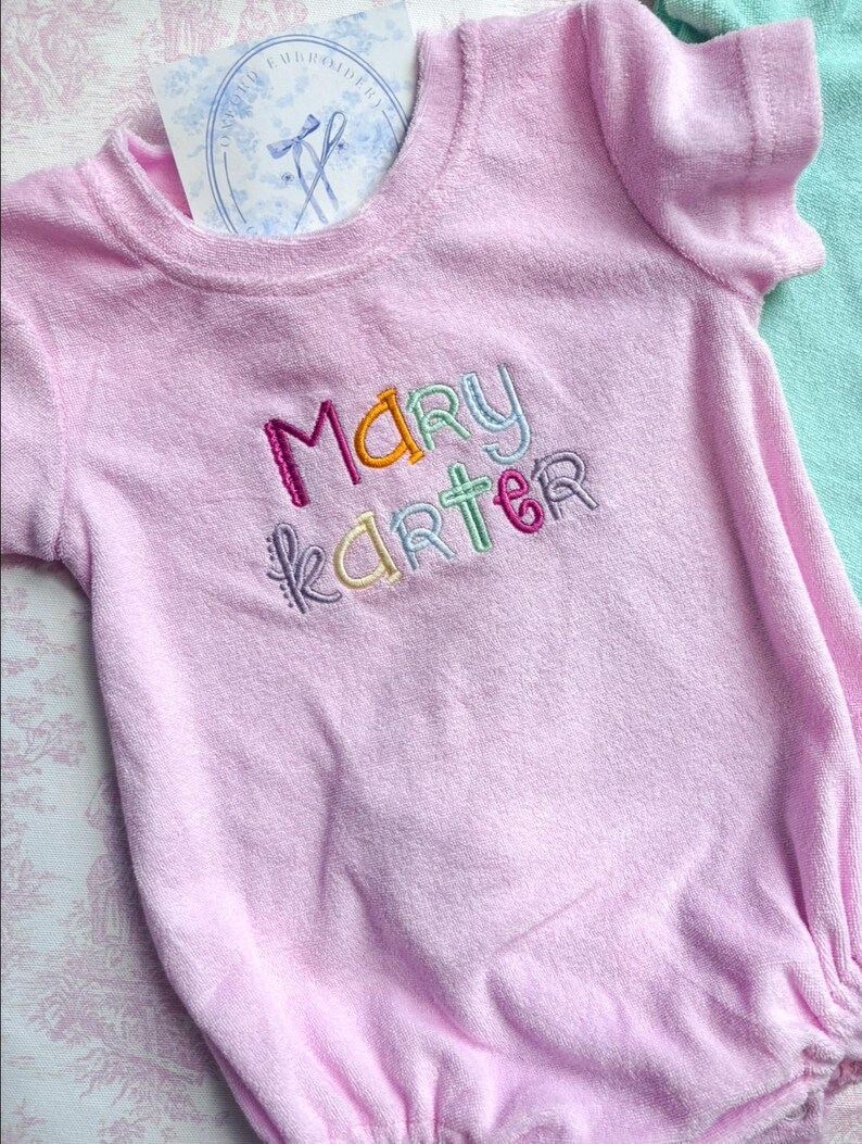 May include: A light pink terry cloth baby romper with the name "Mary Karter" embroidered in colorful thread. The romper has short sleeves and a round neckline. A small card with a logo is tucked into the neckline.