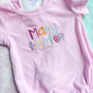 May include: A light pink terry cloth baby romper with the embroidered name "MARY KARTER" in colorful thread. The romper has short sleeves and a round neckline. A card with a logo is tucked into the neckline.