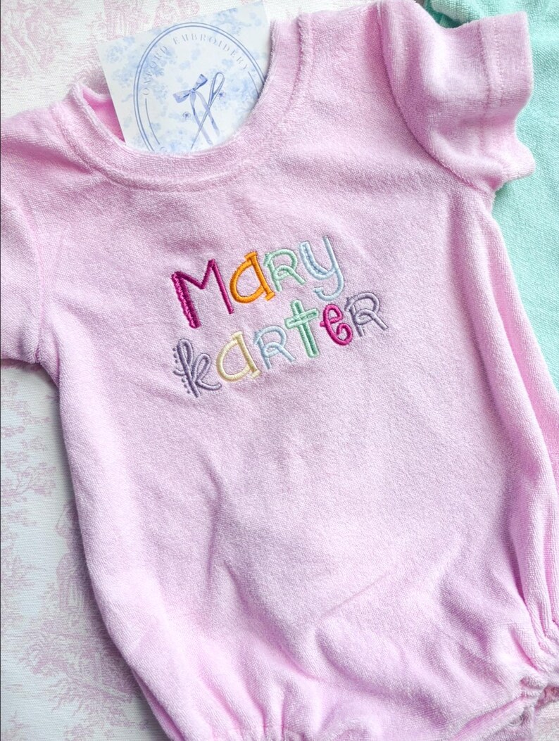 May include: A light pink terry cloth baby romper with the embroidered name "MARY KARTER" in colorful thread. The romper has short sleeves and a round neckline. A card with a logo is tucked into the neckline.