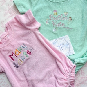 May include: Two baby outfits, one pink and one mint green, with embroidered names and aquatic-themed designs. The pink outfit has the name "Mary Karter" in colorful thread. The green outfit features the name "Brettelee Kate" with a mermaid and fish.