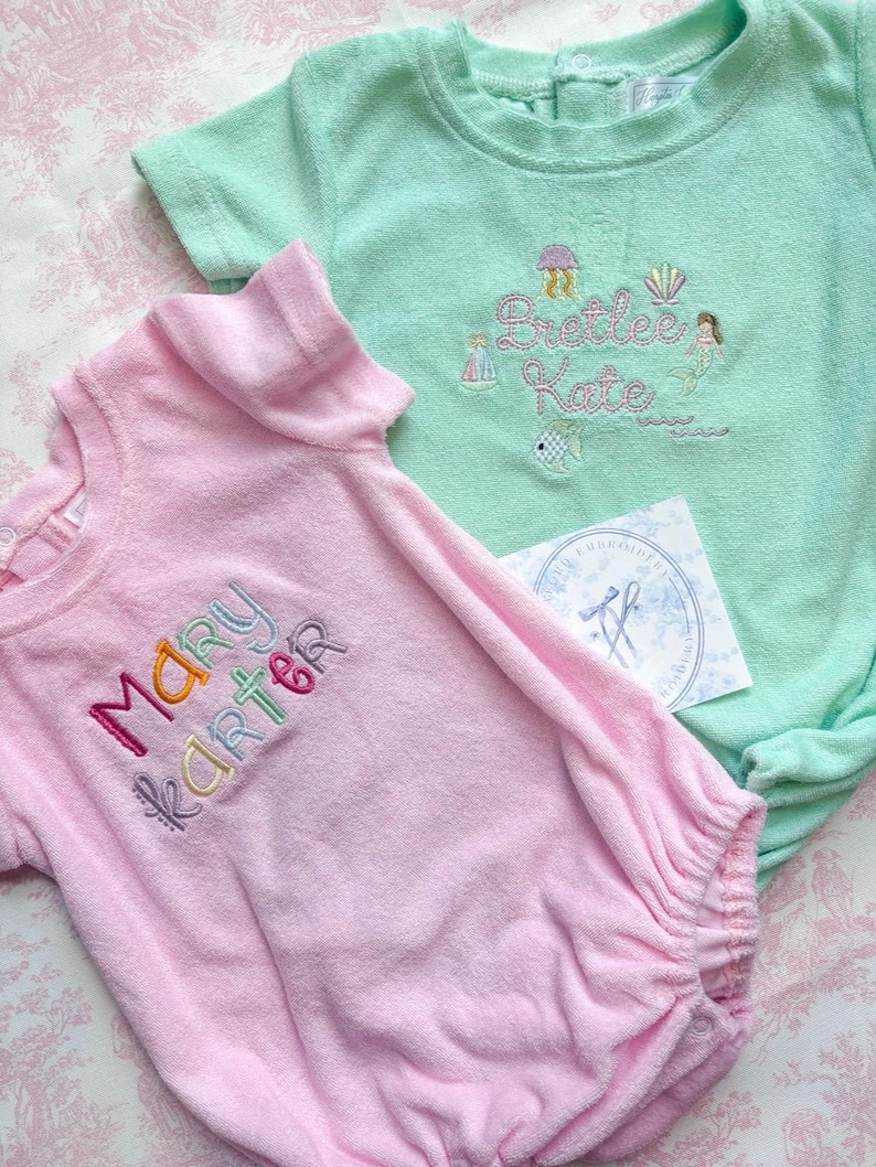 May include: Two baby outfits, one pink and one mint green, with embroidered names and aquatic-themed designs. The pink outfit has the name "Mary Karter" in colorful thread. The green outfit features the name "Brettelee Kate" with a mermaid and fish.