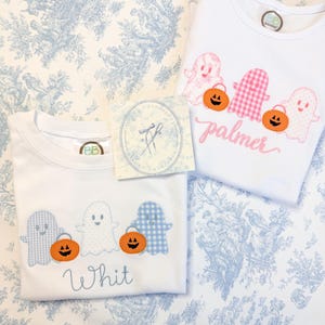 May include: Two white short-sleeved shirts featuring embroidered Halloween designs. One shirt displays blue ghosts and orange pumpkins with the name Whit. The other shirt displays pink ghosts and orange pumpkins with the name palmer.