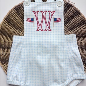 May include: A light blue and white gingham baby romper with a red and blue embroidered monogram and American flag accents. The romper has a button closure and straps.