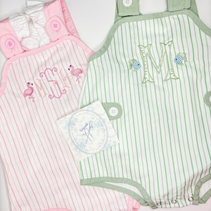 May include: Two baby rompers, one pink and one green, with vertical stripes. The pink romper has a flamingo and monogram embroidery. The green romper features a large embroidered letter and fish. Both have button details and a tag.