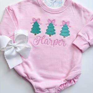 Puede incluir: Romper rosa para bebé con mangas largas. Presenta tres árboles de Navidad turquesa bordados con lazos rosas y el nombre "Harper" en cursiva. Un gran lazo blanco está en el lateral. Una tarjeta con un logotipo está metida en el cuello.