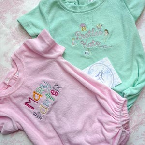 May include: Two terry cloth baby rompers, one light pink and one mint green, embroidered with names and whimsical designs. The pink romper says "Mary Karter" in colorful thread. The green romper says "Preetee Kate" with sea-themed embroidery.