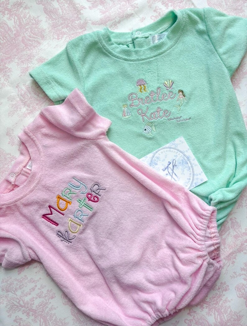 May include: Two terry cloth baby rompers, one light pink and one mint green, embroidered with names and whimsical designs. The pink romper says "Mary Karter" in colorful thread. The green romper says "Preetee Kate" with sea-themed embroidery.
