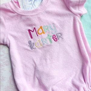 May include: A light pink terry cloth baby romper with the embroidered name "MARY KARTER" in colorful thread. The romper has short sleeves and a round neckline. A tag is tucked into the neckline.