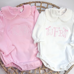 Könnte beinhalten: Zwei rosa und weiße langärmelige Baby-Bodys mit Rüschenkragen. Der rosa Body hat den Namen "LESLIE" auf der Vorderseite gestickt. Der weiße Body hat das Monogramm "JFB" auf der Vorderseite gestickt.